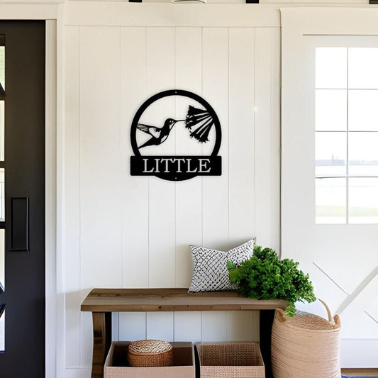 Personalized metal sign that has a hummingbird and a flower in the middle. It is a round metal sign and at the bottom is where you add your wording. It is hanging in a farmhouse entryway above a bench.